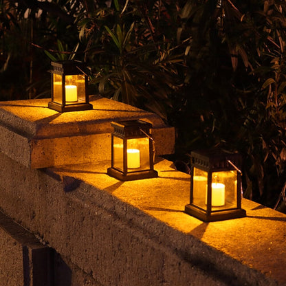 Trois lanternes Lampe solaire de jardin haut de gamme-Classic-Solar™ ornées de bougies scintillantes sont posées au sommet d'un mur de pierre la nuit, diffusant une lumière jaune chaleureuse. Des plantes vertes en arrière-plan créent une ambiance chaleureuse et idéale pour votre jardin.